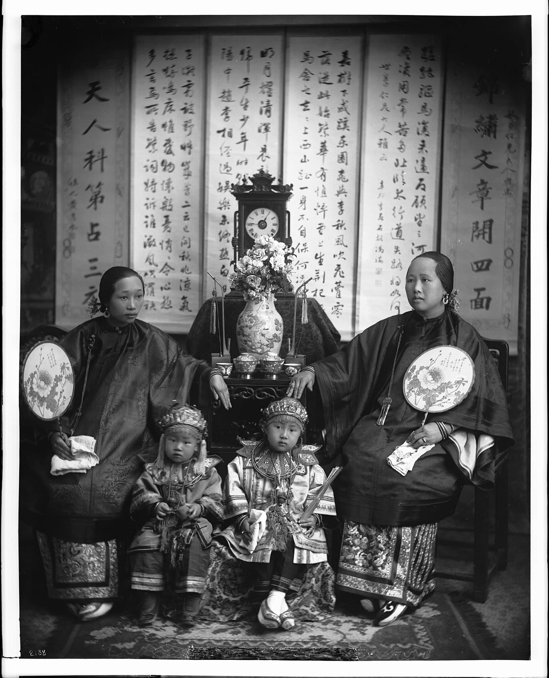A studio portrait from about 1900: two women in embroidered jackets with two small children, calligraphy scrolls behind them