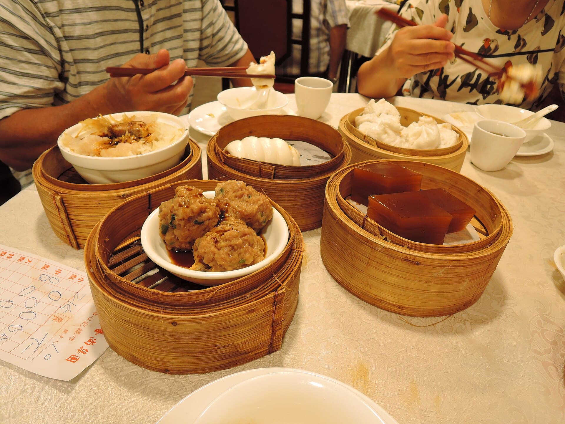 Bamboo steamers of dim sum on a restaurant table, with chopsticks reaching in and a handwritten order card